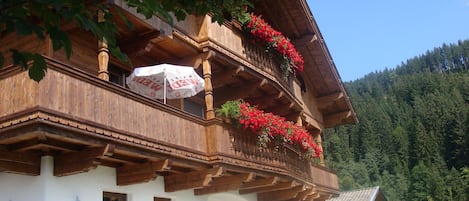 Una cabaña de madera tradicional con balcón decorado con flores rojas, fachada blanca y vistas despejadas de una cadena montañosa boscosa.
