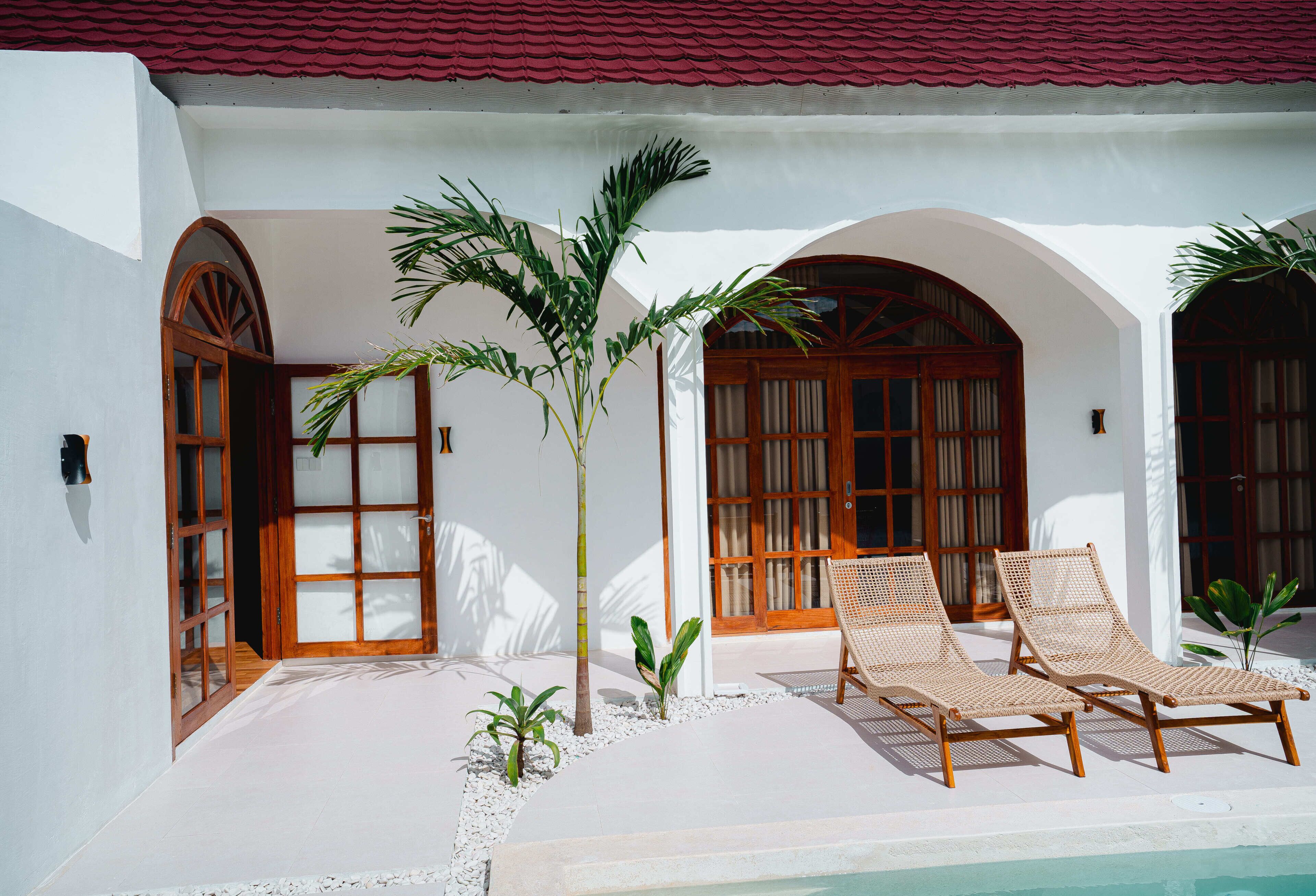 A poolside area with two lounge chairs, a palm tree, and a building with arched doorways.