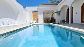 A clear blue swimming pool with white walls and arches, a sun terrace with seating, and a view of the sky.