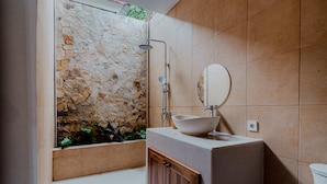 A bathroom with a stone wall, a sink with a round mirror, and a shower. - Costa Luna, Modern Mediterranean Villa Near Lancing Beach (Nusa Tenggara Bar.)