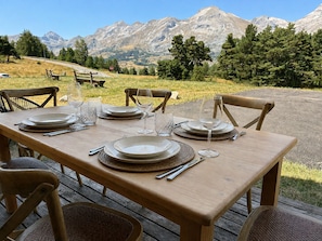 Une table à manger installée à l’extérieur, avec une vue sur les montagnes, des chaises en bois et un banc en arrière-plan.