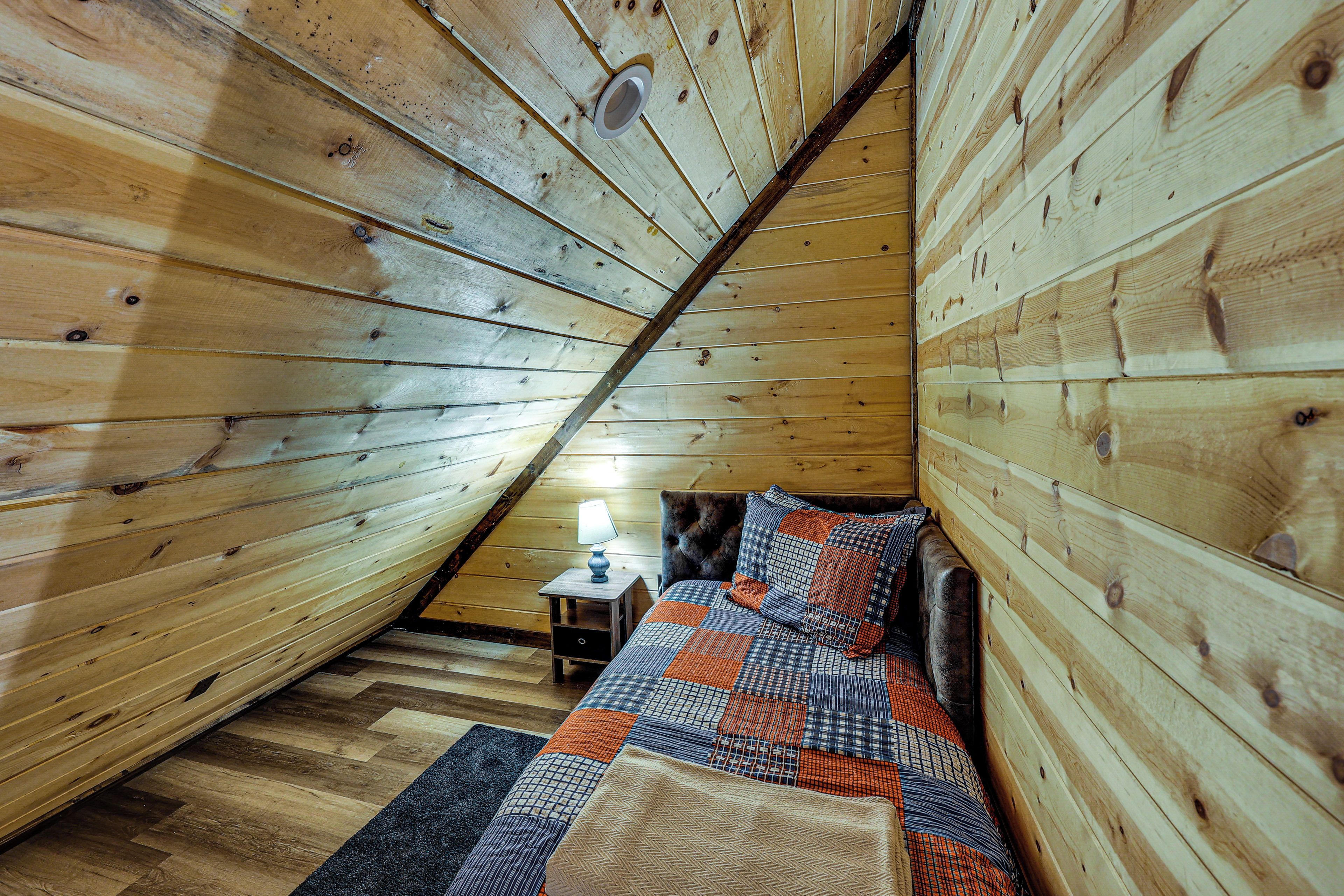 Un dormitorio acogedor en el ático, con techo inclinado, una cama con colcha estampada, una mesita de noche con lámpara y un sillón de cuero oscuro.