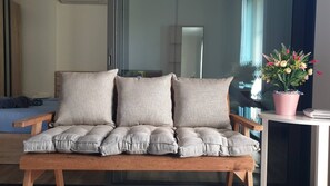 A wooden bench with three cushions, a potted plant with flowers, and a bed with rolled towels in the background.
