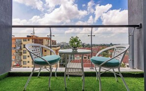Ein Balkon mit zwei weißen Korbstühlen und einem kleinen Tisch mit Blick auf eine Stadtlandschaft, Gebäude und einen klaren Himmel.