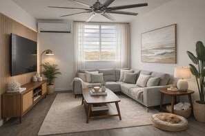 Ein modernes Wohnzimmer mit Flachbildfernseher, Deckenventilator, großem Fenster mit Vorhängen, grauem Sofa, Holz-Couchtisch und kleinem Beistelltisch mit Lampe.