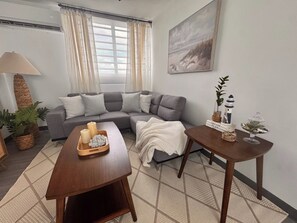 Ein Wohnzimmer mit einem grauen Ecksofa, einem Holztisch mit Kerzen und einer Ablage, einem kleinen runden Tisch mit Dekoration und einem großen Fenster mit Vorhängen.