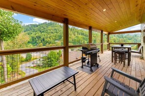 En overdækket terrasse med grill, siddepladser og udsigt til et skovområde.