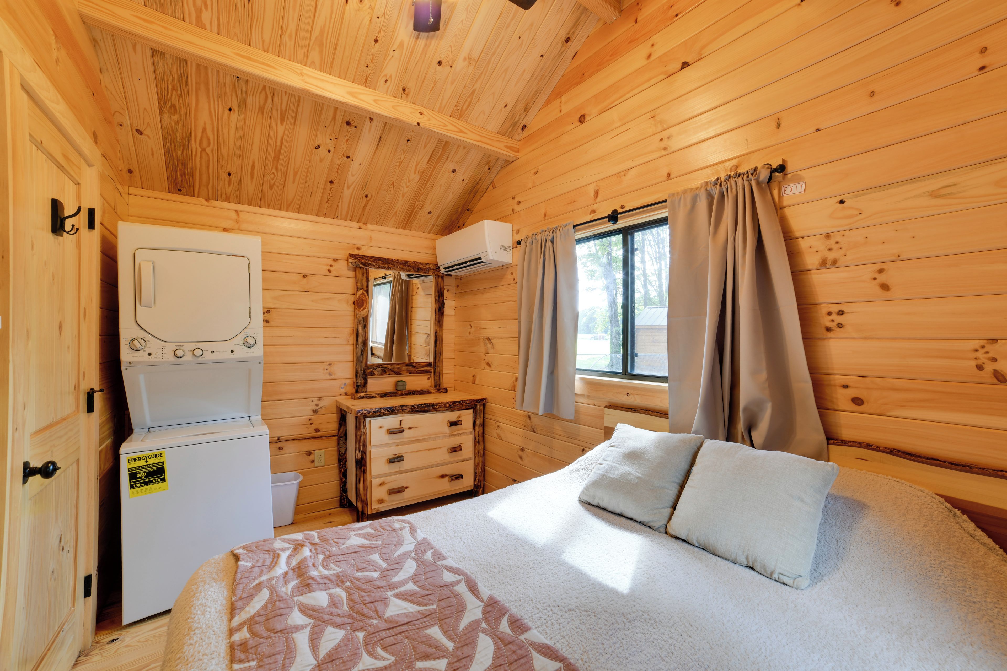 Une pièce de cabane en bois comprenant un lit, une commode, un lave-linge et une fenêtre avec des rideaux.