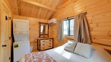 Une pièce de cabane en bois comprenant un lit, une commode, un lave-linge et une fenêtre avec des rideaux.
