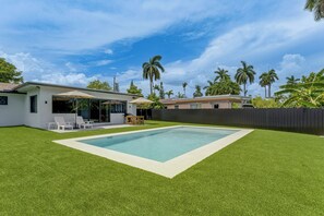 Ein Garten mit Swimmingpool, einer Terrasse mit Stühlen und einem eingezäunten Bereich mit Palmen.