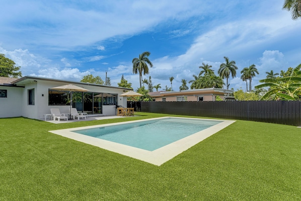 Ein Garten mit Swimmingpool, einer Terrasse mit Stühlen und einem eingezäunten Bereich mit Palmen.