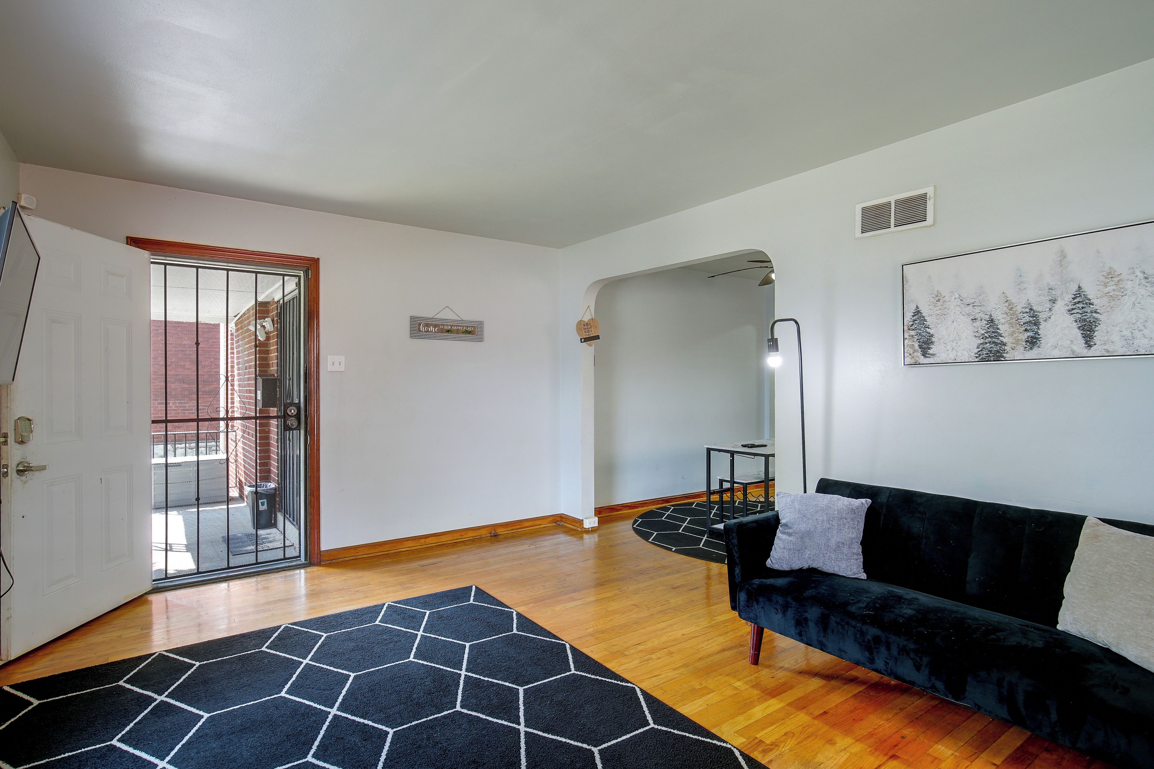 Un salon avec un canapé noir, un tapis à motifs géométriques, un plancher en bois, une porte donnant sur l’extérieur et un mur orné d’un tableau encadré.