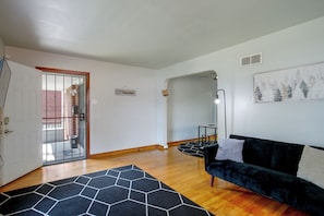 Ein Wohnzimmer mit einer schwarzen Couch, einem geometrischen Teppich, Holzdielen, einer Tür, die nach draußen führt, und einer Wand mit einem gerahmten Bild.