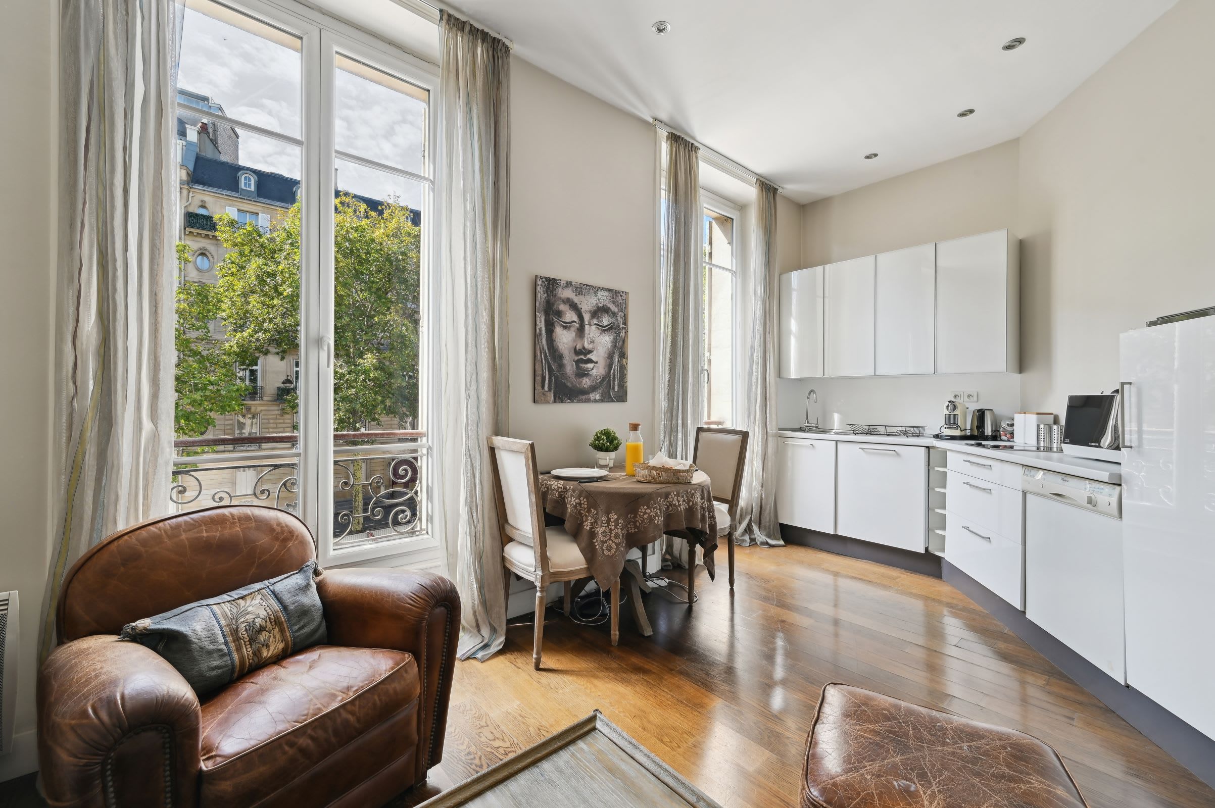 A modern kitchen with white cabinets, a dining table with chairs, and a view of a building through large windows.