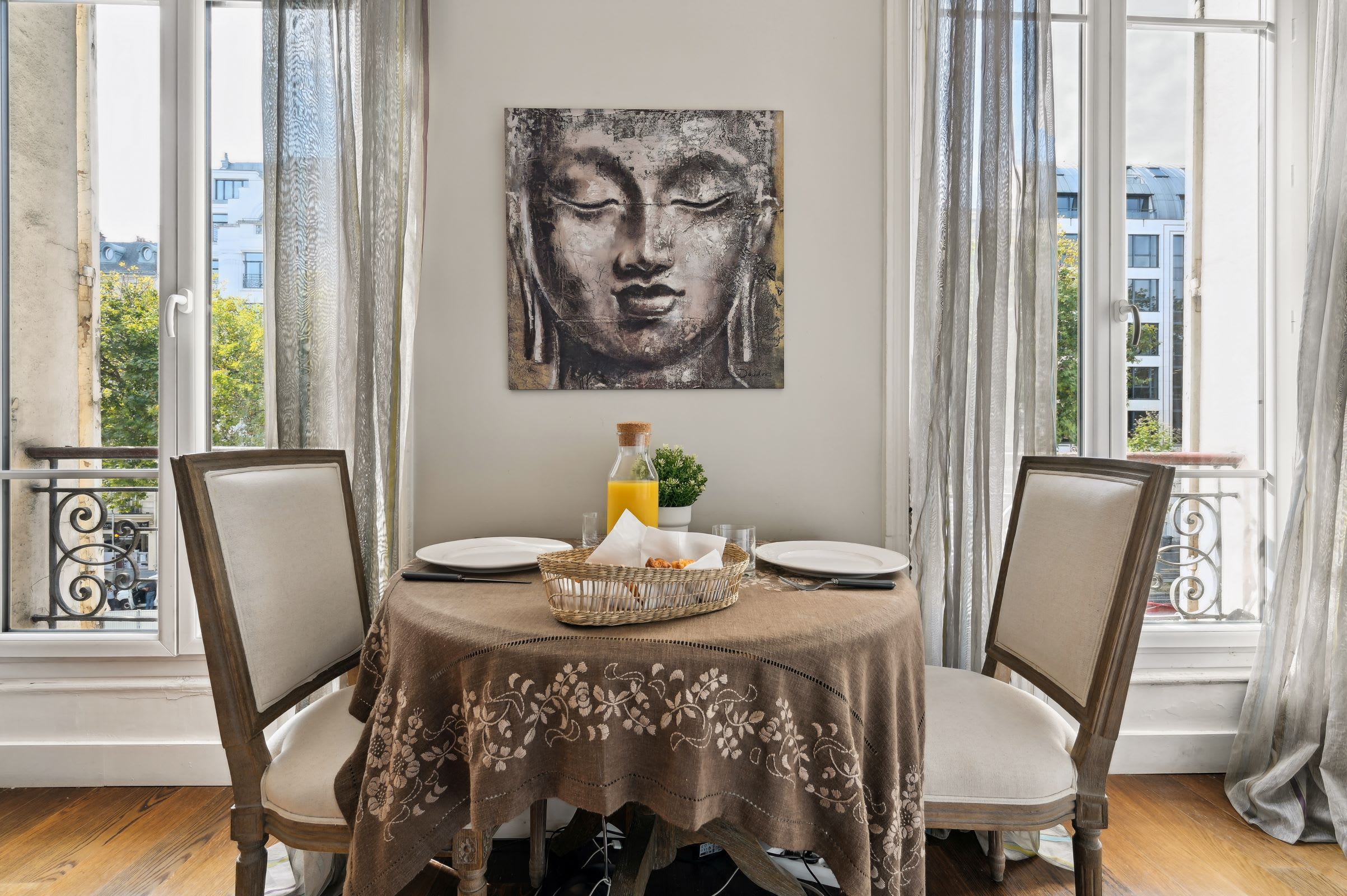 Un coin repas avec une table dressée pour deux personnes, un tableau représentant un Bouddha accroché au mur et une vue sur les bâtiments à travers de grandes fenêtres.