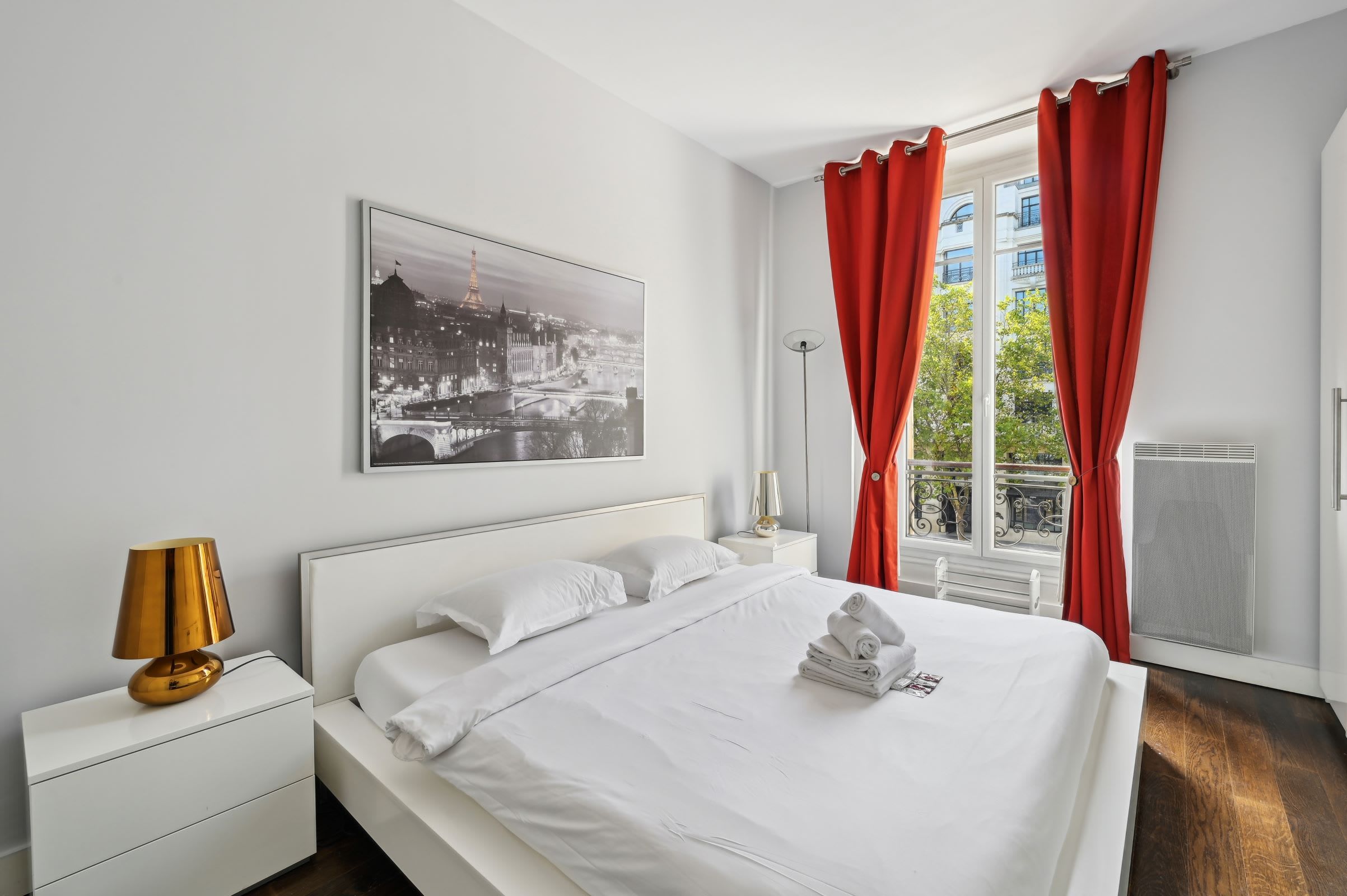 A hotel room with a bed, white nightstands, a lamp, a window with red curtains, and a cityscape view.