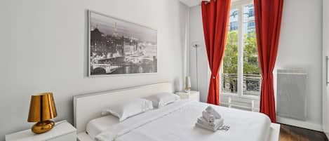A hotel room with a bed, white nightstands, a lamp, a window with red curtains, and a cityscape view.