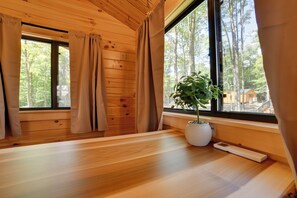 L'intérieur d'une cabane avec des murs en bois, une grande fenêtre avec des rideaux, une plante en pot et un thermostat placé sur le rebord de la fenêtre.