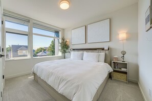 Une chambre à coucher avec un grand lit, deux fenêtres, une table de chevet avec une lampe et une œuvre d’art accrochée au mur.