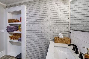 A bathroom with a white bathtub, a white sink, and a white vanity. There is a white brick wall and a white door leading to a bathroom storage area with shelves. - Downtown Salem Home: Walk to Main Street! (Salem)