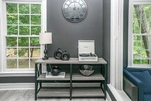 Quarto com parede cinzenta, relógio redondo, mesa com abajur, alto-falante e cesta.