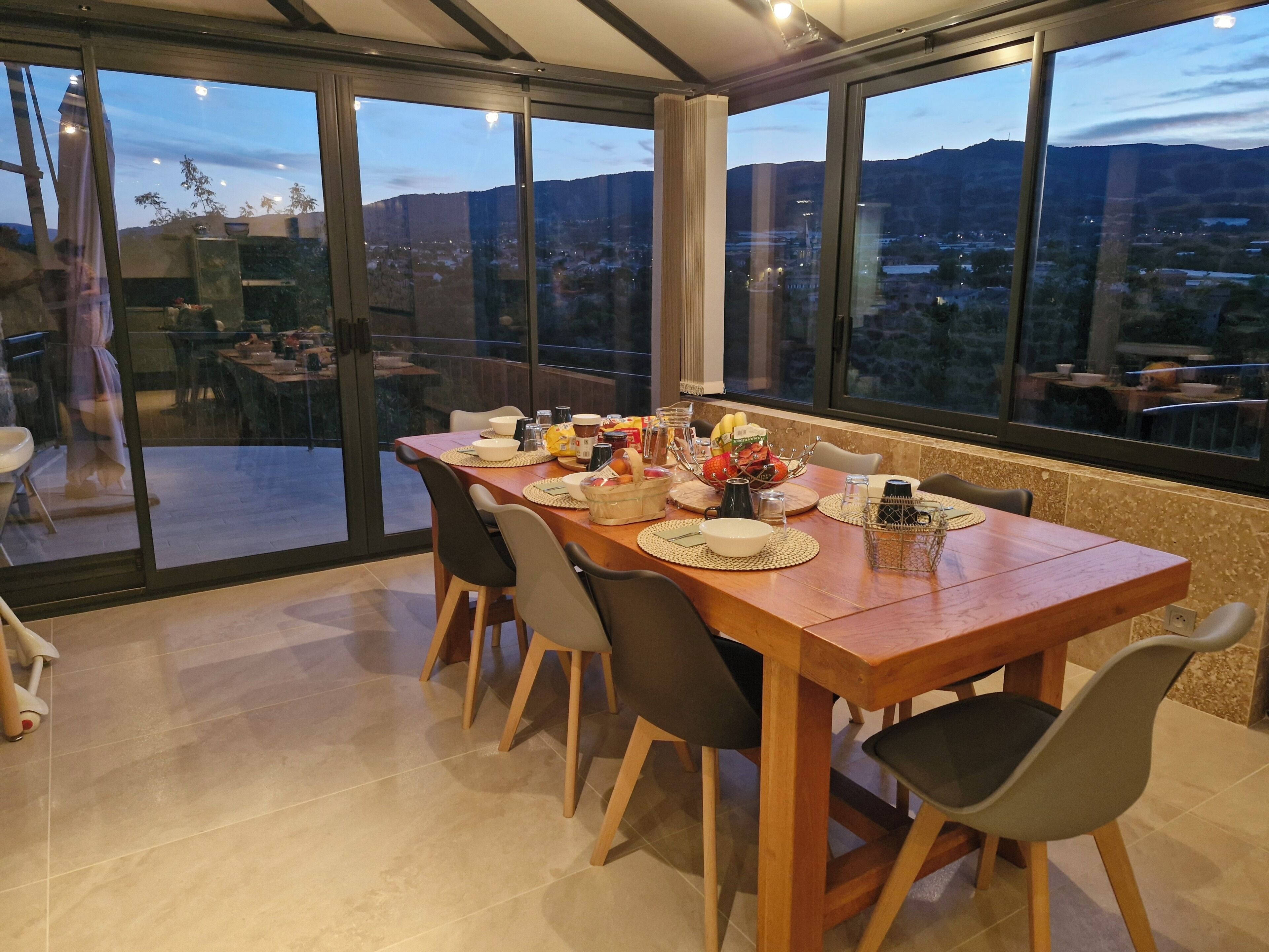 A dining area with a large table set for four, surrounded by chairs, with a view of a cityscape and mountains through large windows.