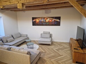Un salon doté d’un parquet en bois à motifs chevrons, d’un canapé gris, d’un fauteuil gris plus petit, d’une table basse ronde et d’un téléviseur à écran plat posé sur un meuble en bois.