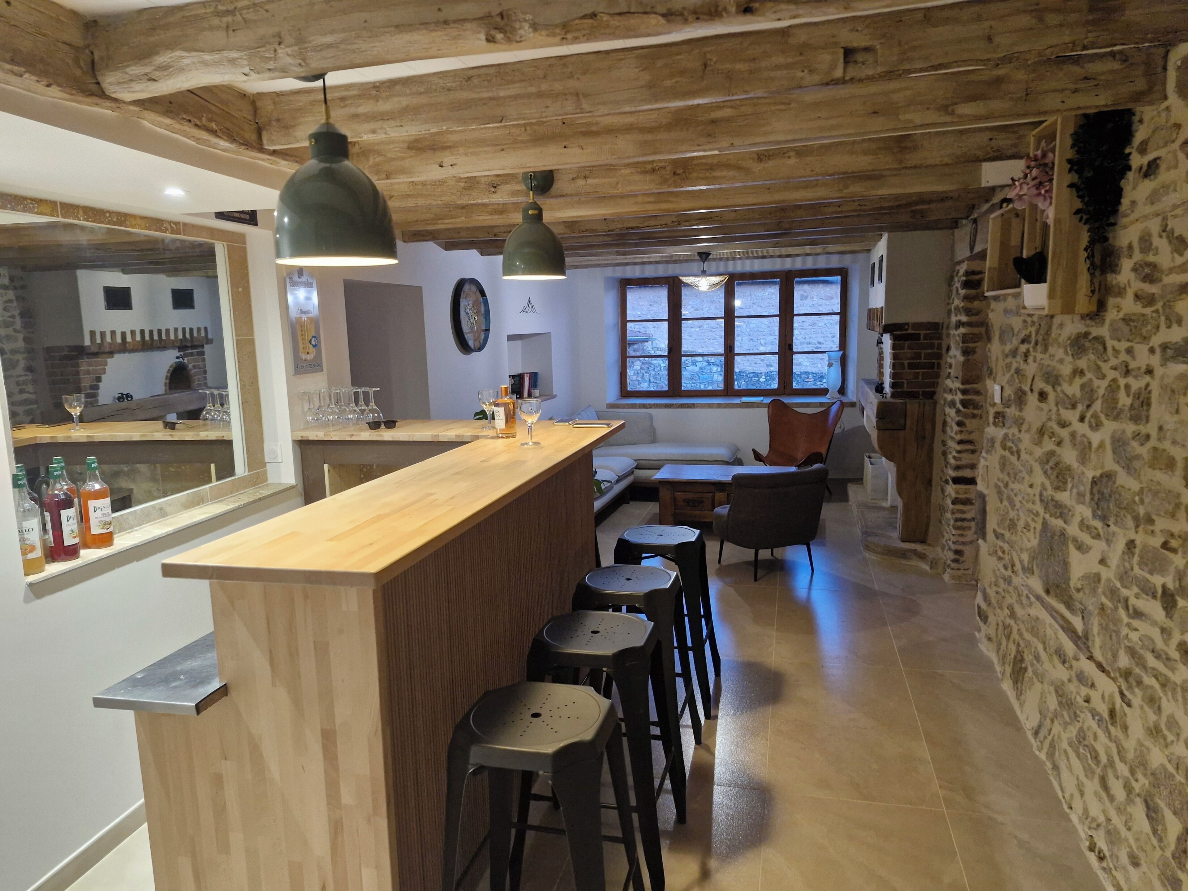 A rustic kitchen with a wooden bar and stools, a stone wall, and a large window with a view.