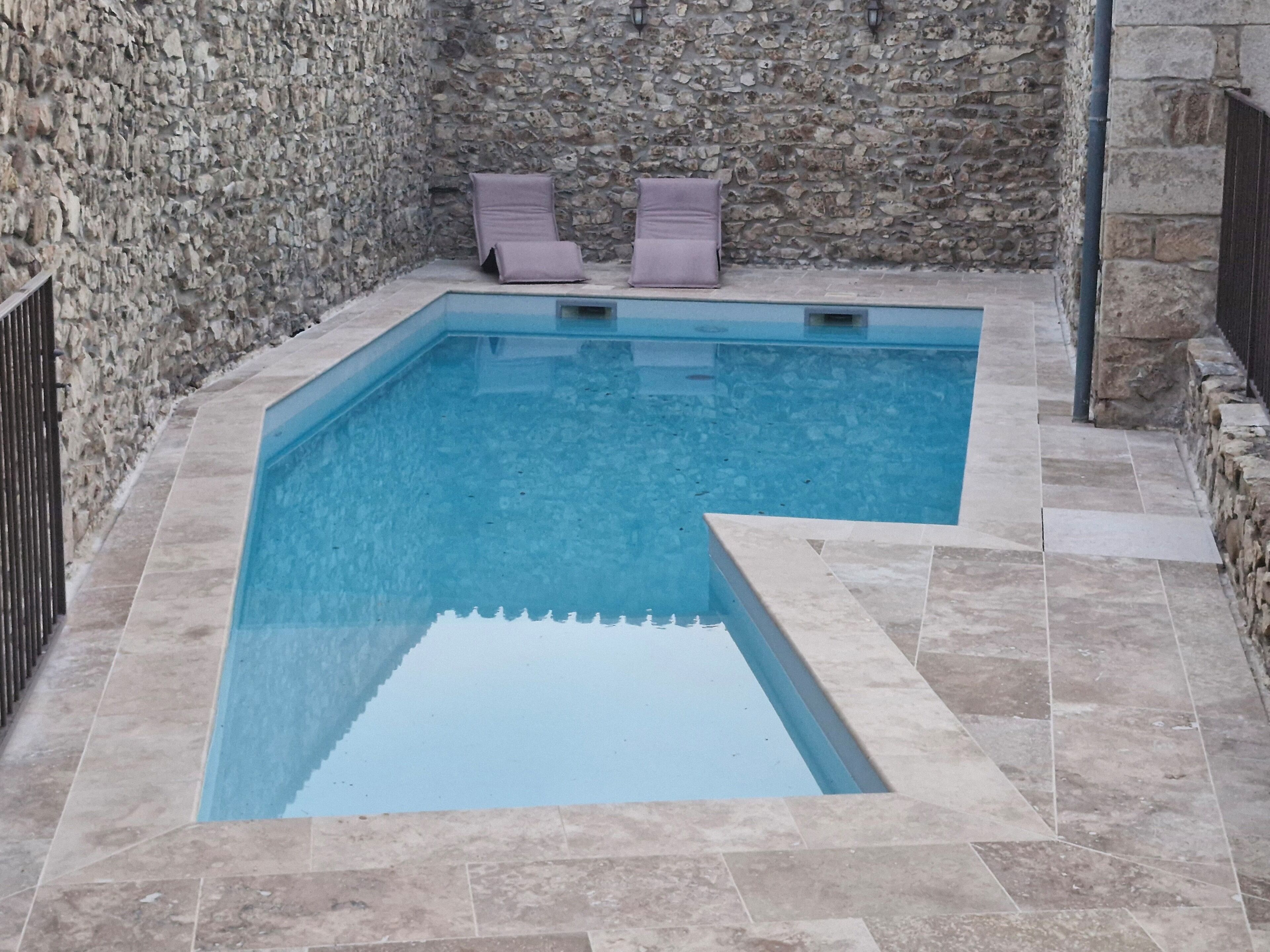 A rectangular swimming pool with a stone wall and tiled floor, two lounge chairs, and a stone wall.