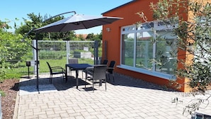 Eine Terrasse im Freien mit Tisch und Stühlen, einem schattenspendenden Sonnenschirm und einem Gebäude mit großen Fenstern.