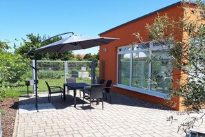 Een terras buiten met een tafel en stoelen, een overdekte parasol en een gebouw met grote ramen.