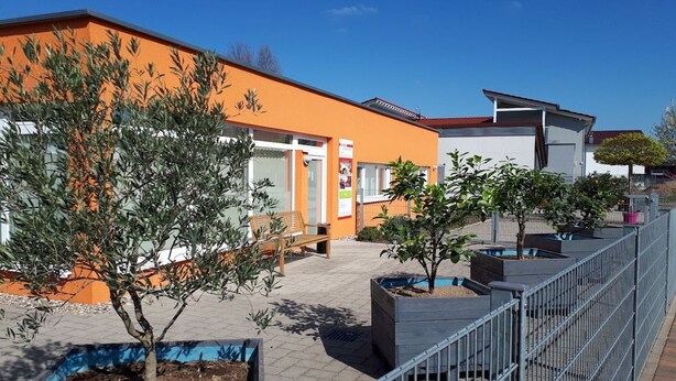 Ein Gebäude mit einer orangefarbenen Fassade, ein von einer Hecke umgebener Außenbereich mit Blumentöpfen und ein klarer Himmel.