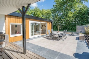 Un patio exterior moderno con mesa y sillas, una terraza de madera y un ventanal que da a un espacio interior.