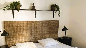 En säng med vita sängkläder, två kuddar, en vävd sänggavel, sänglampor och en hylla med växter.