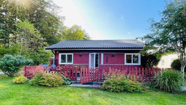 Ett rött trähus med ett tak täckt av tegelpannor, omgivet av ett trästaket och frodig grönska.