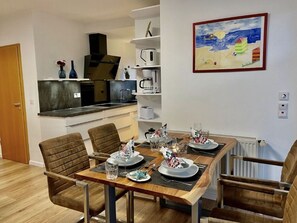 Un comedor con una mesa de madera dispuesta para cuatro, una cocina al fondo y un cuadro en la pared.