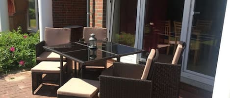An outdoor patio with a glass table and wicker chairs, a brick wall, and a sliding glass door.