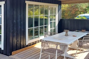 Une terrasse avec une table blanche et des chaises en osier, une grande fenêtre et un mur extérieur de couleur sombre.