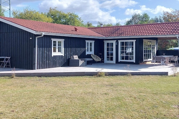 Un bâtiment à deux étages, doté d’un toit recouvert de tuiles rouges, d’un patio abrité avec des meubles d’extérieur et d’une cour gazonnée.