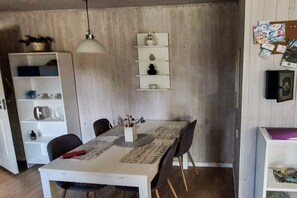 Un coin repas comprenant une table, des chaises, un luminaire suspendu, un meuble blanc avec des étagères et un tableau d’affichage avec des photos.