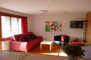Een woonkamer met een rode bank, een zwarte fauteuil, een klein tafeltje en een televisie aan de muur.