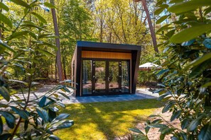 A modern outdoor structure with a glass door, surrounded by a well-maintained garden and trees. - Heideborg Cottage With Terrace (Ruinen)