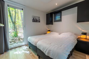 A bedroom with a bed, a nightstand, a wall-mounted lamp, a window with blinds, and a cityscape painting. - Heideborg Cottage With Terrace (Ruinen)