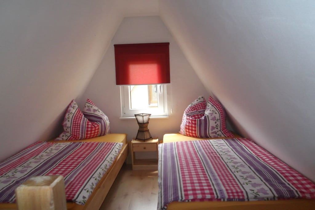 A small, cozy room with two beds, a window with a red blind, and a bedside table with a lamp.