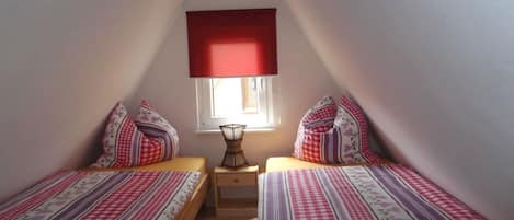 Una habitación pequeña y acogedora con dos camas, una ventana con persiana roja y una mesita de noche con lámpara.