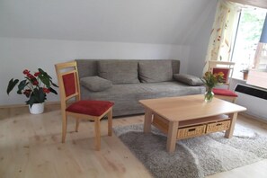 Ein Wohnzimmer mit einem grauen Sofa, einem Holzstuhl mit rotem Kissen, einem Holztisch, einer kleinen Topfpflanze und einem Fenster mit Vorhängen.
