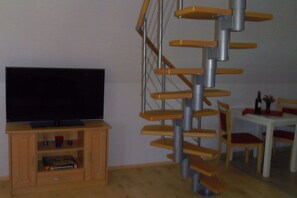 Ein Wohnzimmer mit Fernseher auf einem Holzboden, einer Wendeltreppe und einem Essbereich mit Stühlen und Tisch.