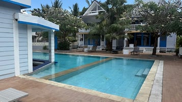En swimmingpool med solstolar och ett överbyggt område, omgiven av palmer och en byggnad med blå detaljer.