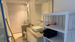 Ein Badezimmer mit einem weißen Waschbecken, einem Spiegel und Regalen; ein Duschbereich mit einer Glastür; und eine Toilette.
