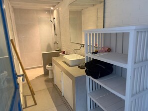 Ein Badezimmer mit einem weißen Waschbecken, einem Spiegel und Regalen; ein Duschbereich mit einer Glastür; und eine Toilette.